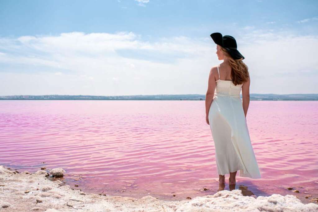 Back view of cute teenager woman wearing white dress and hat walking on a amazing pink lake of sea water and blue sky