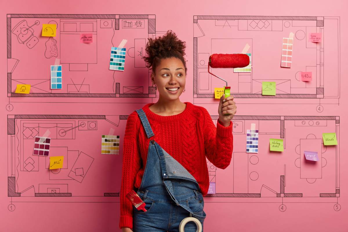 Glad smiling Afro American decorator holds paint roller, busy doing repairing according to design project, paints room red, wears knitted sweater and denim overalls, uses tools for repair and painting