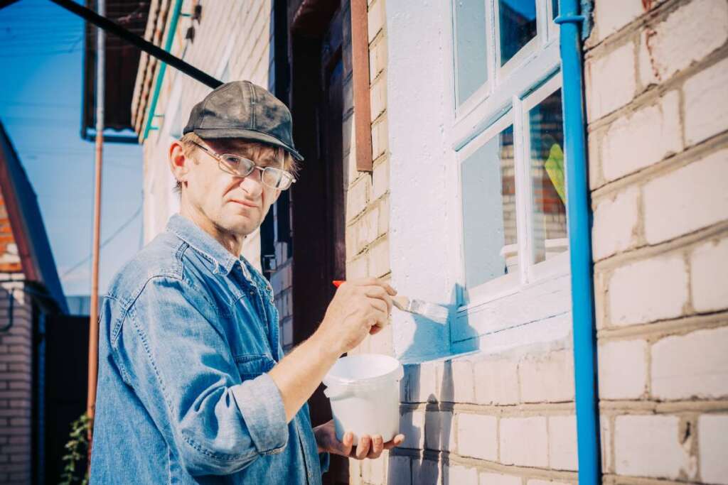 Caucasian Mature Man In Glasses And Cap Painting The Window Frame Outside Of The Private Brick House  In Sunny Day.