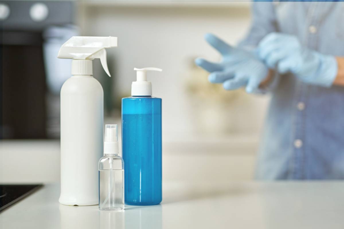 Sanitizer bottles to clean surfaces in the kitchen. Woman putting on protective gloves while getting ready for clean up in the background. Antiseptic, disinfection, cleanliness and healthcare