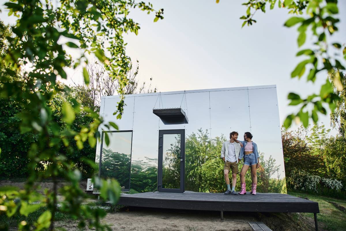 full length of happy and stylish interracial couple standing on porch of glass house in countryside