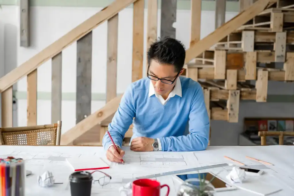 An adult man sits at a brightly lit, modern office table, working intently on blueprints. Wearing a light blue sweater and glasses, he's focused on his designs.