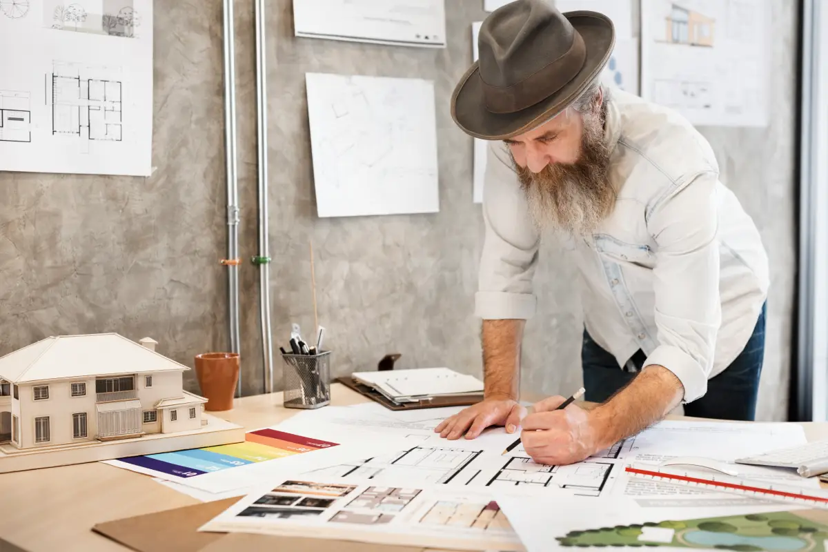 A man with a beard and hat reviews architectural designs on a table in a workspace.